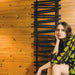 Carisa Vivek Aluminium Towel Radiator fixed to a wooden wall next to a model sitting in a green and black pattern dress