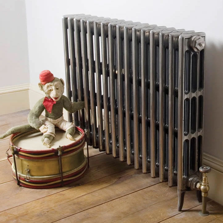 810mm Carron Victorian 4 Column Cast Iron Radiator hand burnished finish in a lifestyle image within hallway with a wooden floor and monkey toy quite dishevelled sitting on an old drum